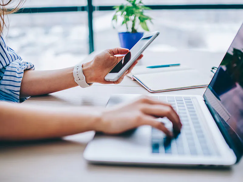 Frau arbeitet an einem Laptop und hält ihr Smartphone in der linken Hand.